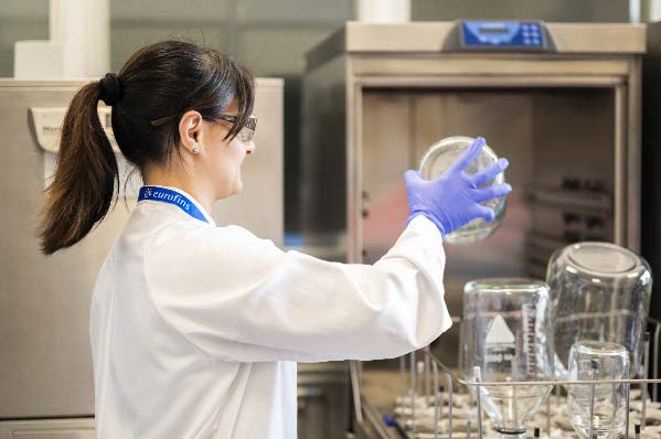 Technicien de laboratoire en blouse blanche et gants bleus manipulant un grand récipient en verre, en cours de chargement ou de déchargement d’un laveur ou stérilisateur de laboratoire. L’image illustre les procédures de nettoyage et de stérilisation des équipements mises en œuvre par Eurofins BioPharma Product Testing pour garantir l’efficacité du cycle de lavage et la conformité aux normes pharmaceutiques.