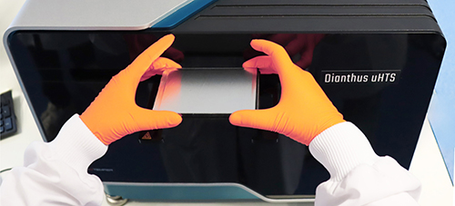 Mains gantées en orange manipulant un support transparent devant un appareil Dianthus uHTS dans un laboratoire Eurofins. L’image illustre une opération de préparation ou d’analyse d’échantillons dans le cadre de criblage à haut débit (HTS), mettant en avant l’utilisation de technologies avancées pour la recherche scientifique, la bioanalyse et le développement pharmaceutique.