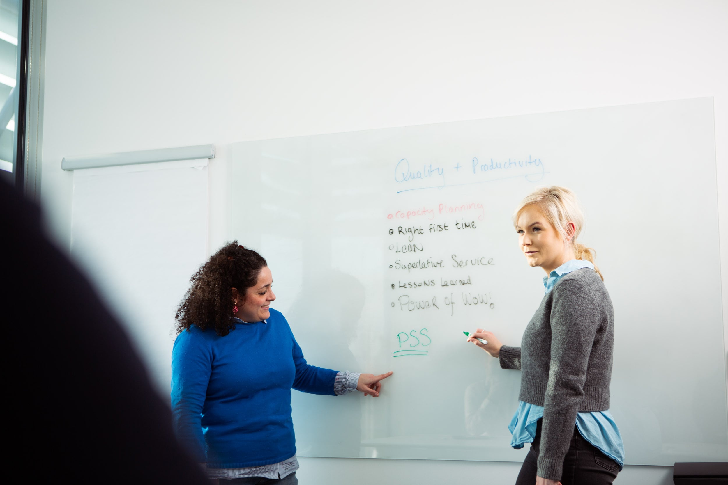 Session de formation ou réunion de travail chez Eurofins BioPharma Product Testing, avec deux personnes debout devant un tableau blanc. Le tableau contient des notes écrites à la main sur la qualité et la productivité, incluant des points tels que “Right First Time”, “Clean”, “Superlative Service”, et “Lessons Learned”. Une personne tient un marqueur et l’autre pointe vers le tableau, illustrant une discussion active sur les processus et l’amélioration continue.