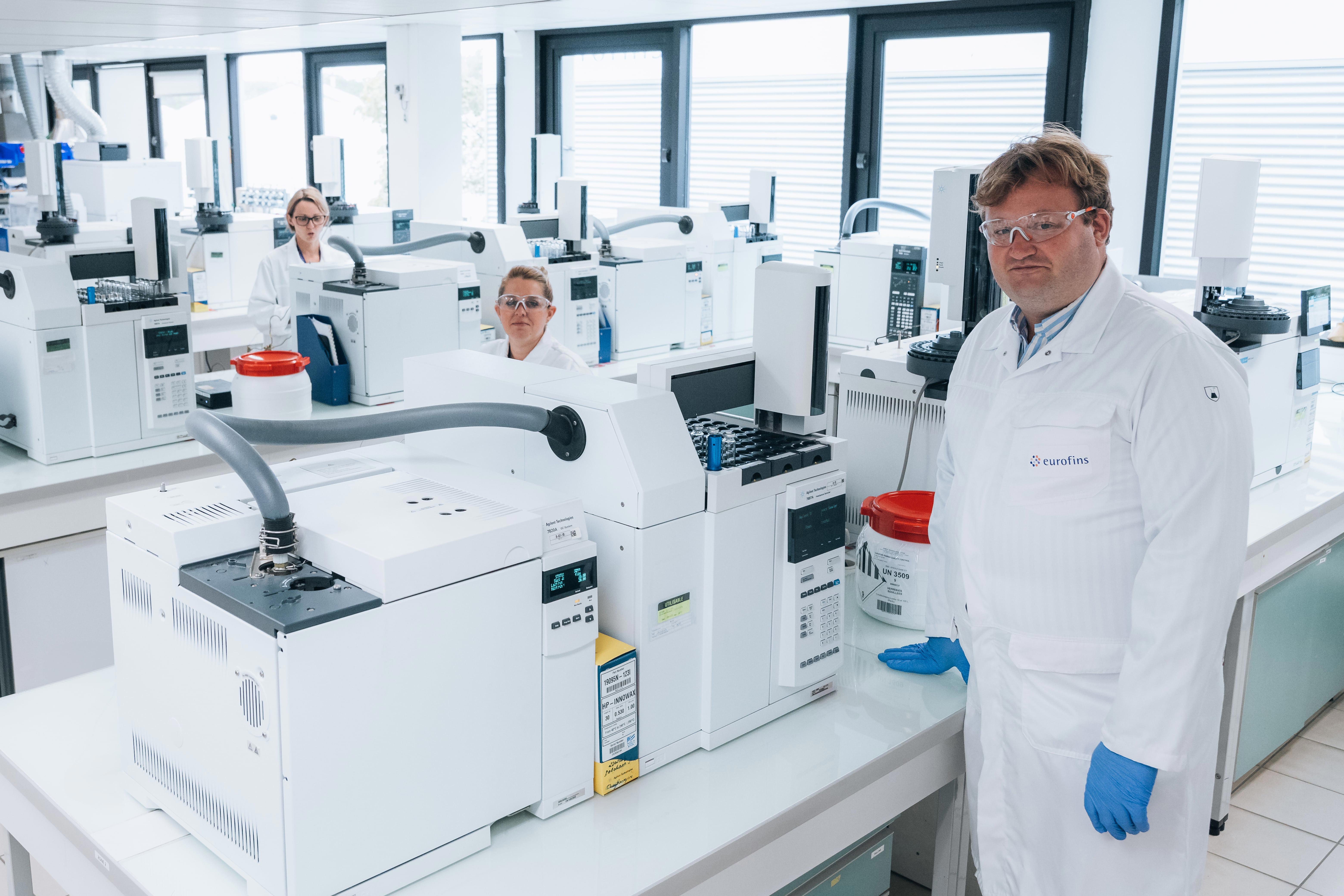 Laboratoire moderne d’Eurofins BioPharma Product Testing, où plusieurs scientifiques en blouses blanches travaillent sur des équipements de chromatographie en phase gazeuse. Au premier plan, une personne équipée de lunettes de protection et de gants bleus manipule un appareil. En arrière-plan, deux autres collaborateurs sont assis devant des instruments similaires. Le laboratoire est lumineux, éclairé par la lumière naturelle provenant de grandes fenêtres.
