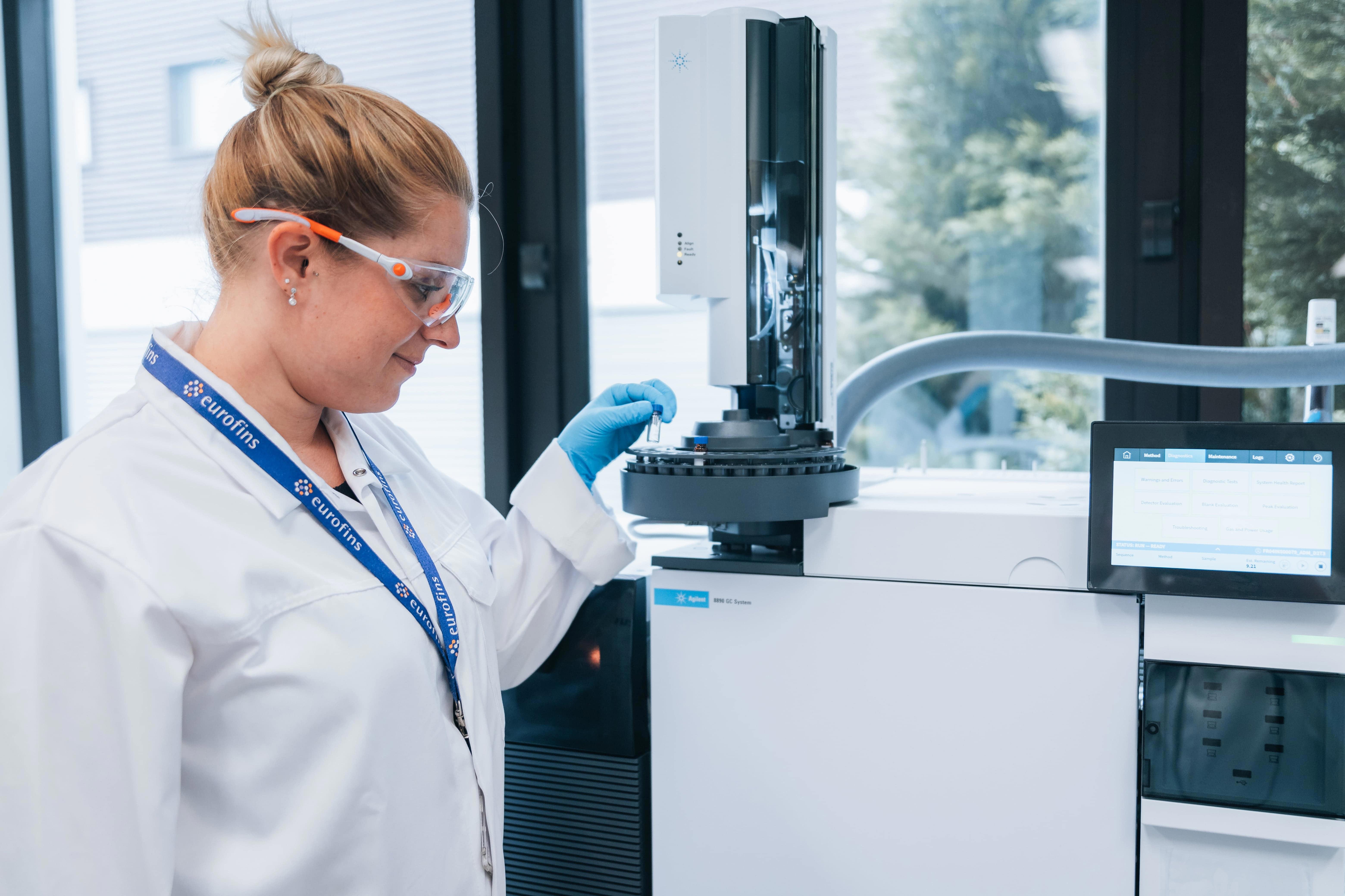 Technicienne de laboratoire portant une blouse blanche, des lunettes de protection, des gants nitrile bleus et un badge Eurofins, en train de manipuler un instrument analytique équipé d’un écran de contrôle. L’image illustre les activités de contrôle des résidus de stérilisation réalisées par Eurofins BioPharma Product Testing dans un environnement de laboratoire conforme aux standards pharmaceutiques.