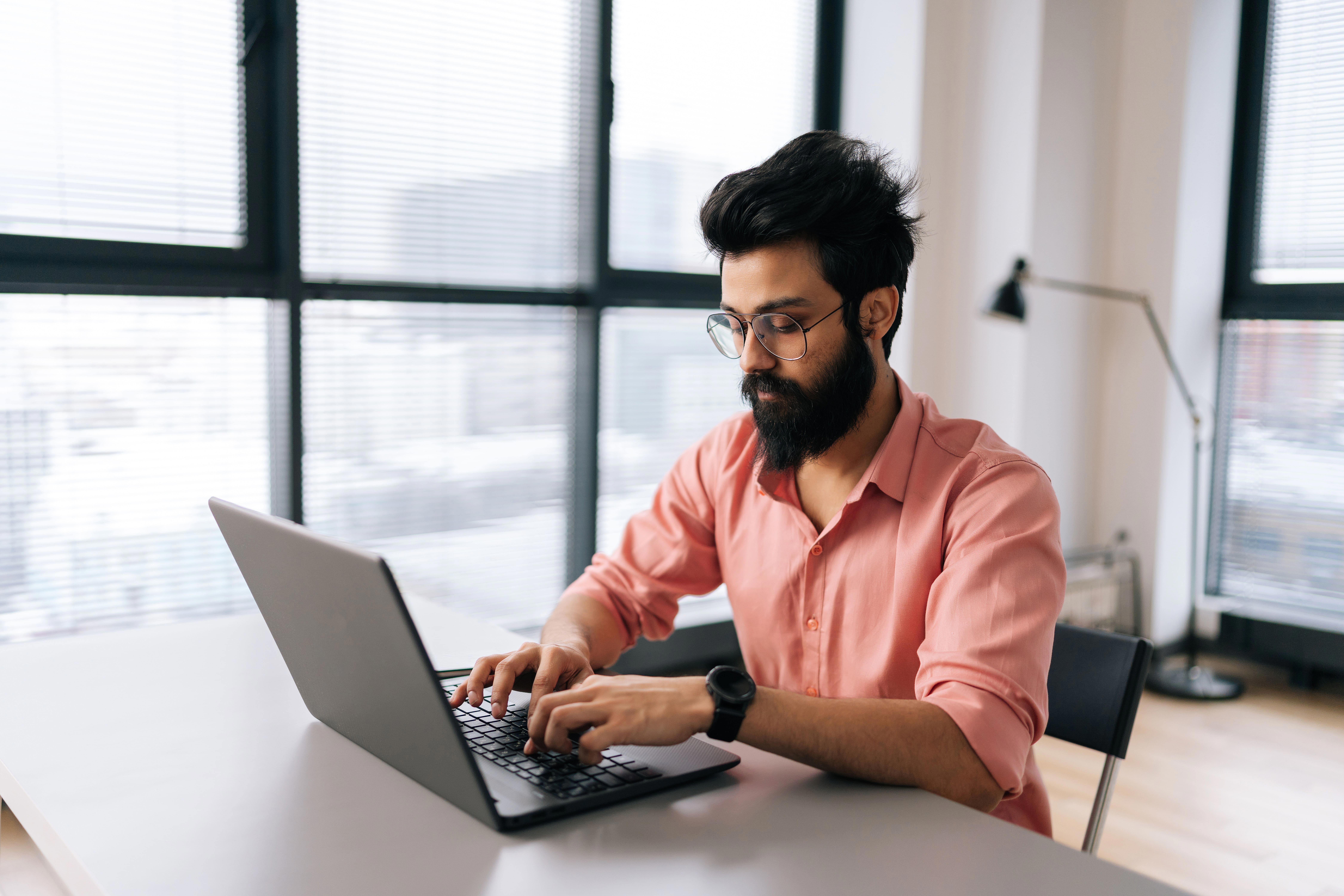 indian-designer-male-in-stylish-eyeglasses-working-on-laptop-sitting-at-desk-in-light-coworking-office-room-by-window-bearded-business-man-wearing-casual-clothes-looking-at-computer-screen_9000-x-6000-px-1-2
