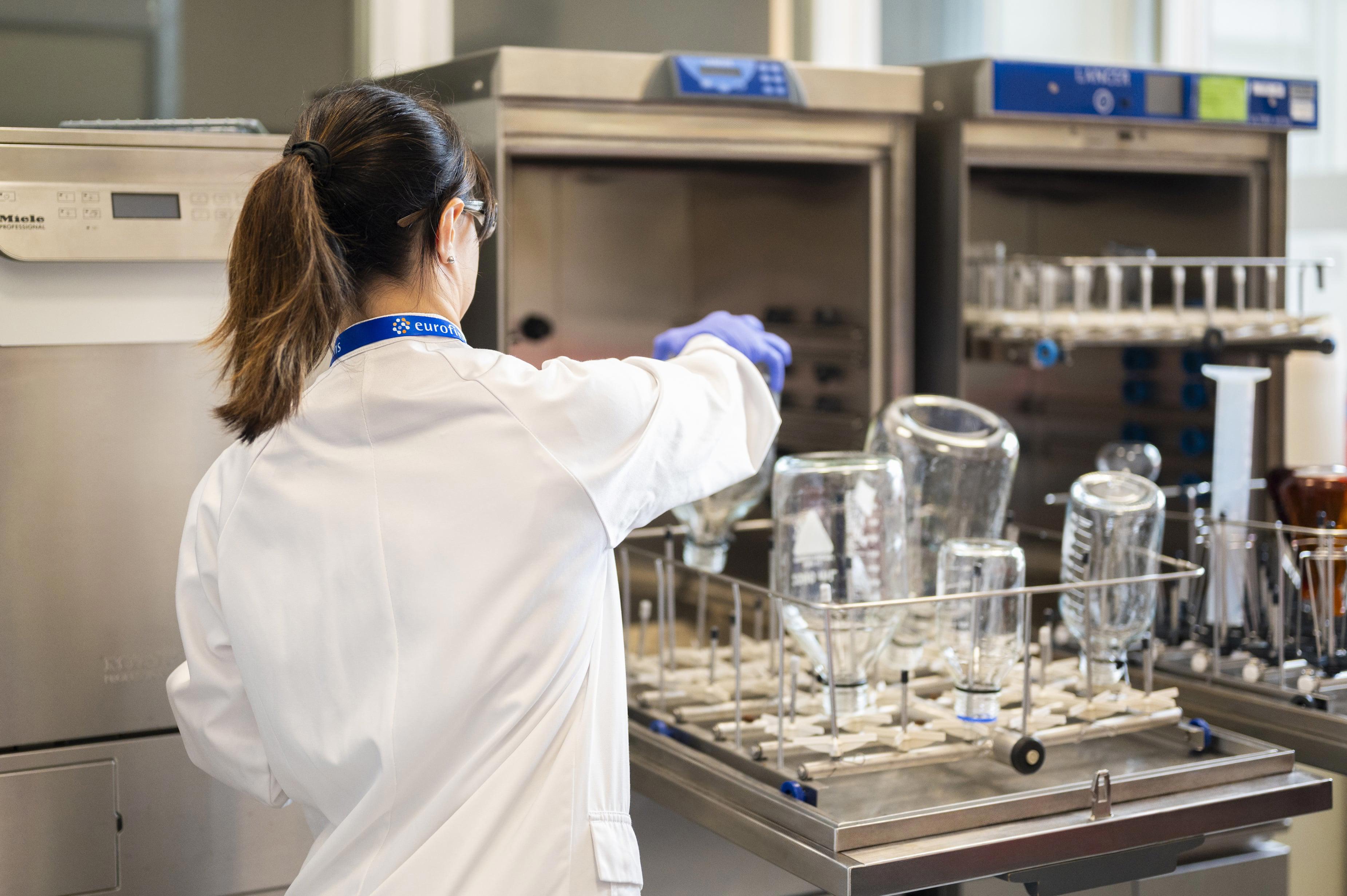 Technicien de laboratoire en blouse blanche et gants violets, manipulant des verreries telles que des béchers et flacons dans un lave-vaisselle de laboratoire. L’image illustre les étapes de validation des équipements de lavage mises en œuvre par Eurofins BioPharma Product Testing pour garantir la propreté et la conformité des matériels utilisés en environnement pharmaceutique.
