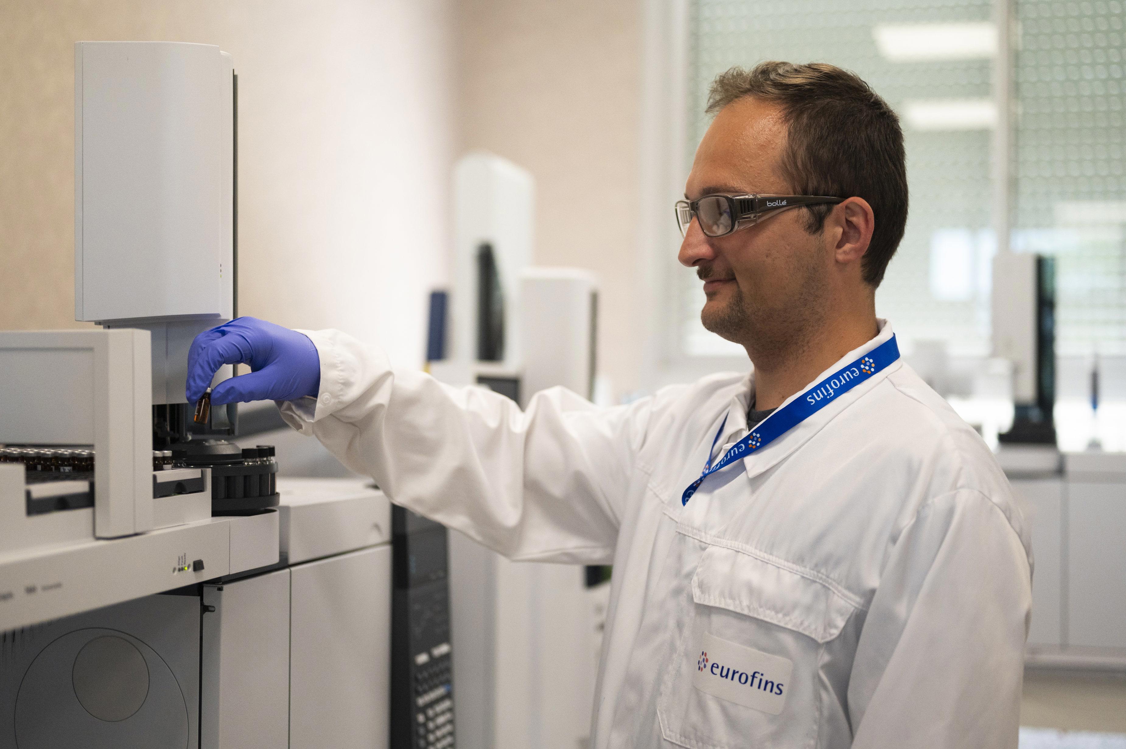 Technicien en blouse de laboratoire Eurofins et gants de protection violets, insérant un flacon d’échantillon dans un chromatographe en phase gazeuse couplé à un spectromètre de masse (GC-MS). L’équipement analytique est composé d’un plateau d’injection automatisé et d’un module principal blanc, utilisé pour l’analyse de composés volatils dans un environnement de laboratoire.