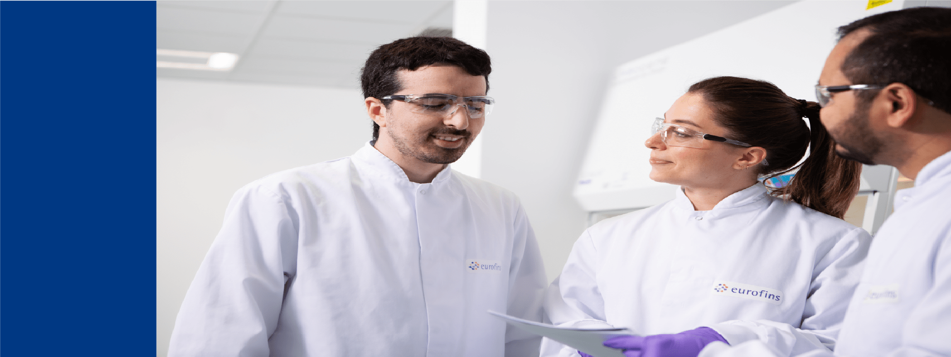 Équipe de laboratoire Eurofins Biopharma Product Testing en discussion devant une hotte de sécurité. Les personnes portent des blouses Eurofins et des gants de protection, tenant des documents dans un environnement scientifique moderne.