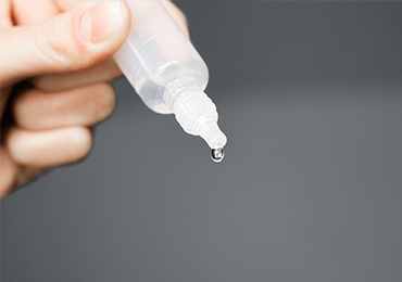 Main tenant un flacon compte-gouttes transparent, avec une goutte de solution en formation à l’extrémité. L’image illustre les formulations liquides destinées à une administration orale ou auriculaire, telles que les solutions buvables, les suspensions ou les gouttes auriculaires. Elle met en lumière les exigences de précision dans le dosage et la manipulation de ces formes, dans le cadre des essais de qualité, de stabilité et de conformité réglementaire réalisés par Eurofins BioPharma Product Testing