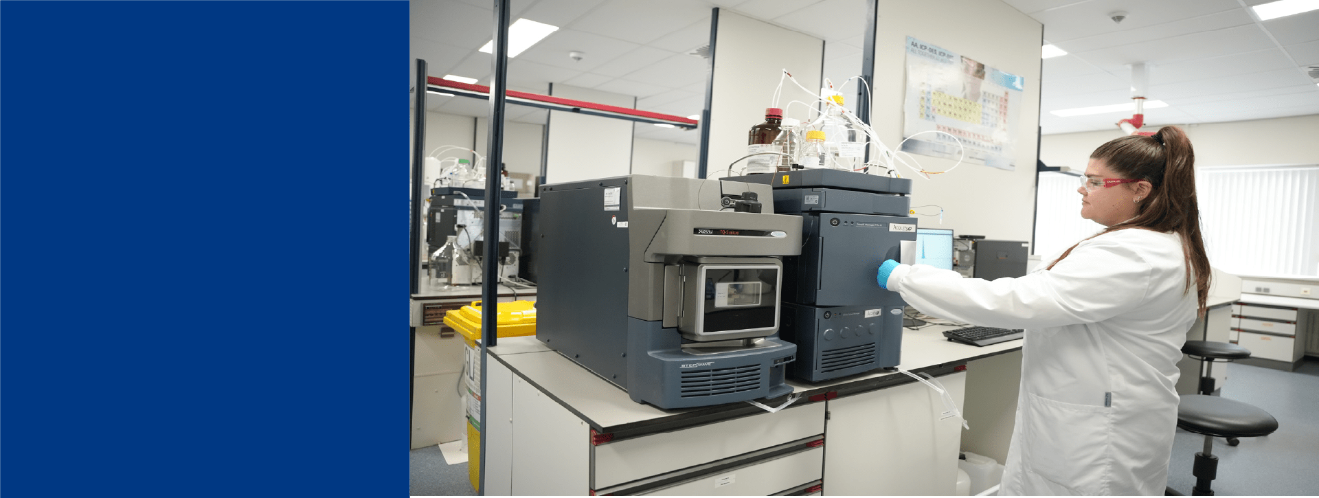 Technicienne de laboratoire Eurofins Biopharma Product Testing travaillant sur un système analytique de chromatographie liquide installé sur une paillasse. L’environnement montre un laboratoire moderne avec des équipements scientifiques, des flacons de réactifs, un ordinateur et des affiches techniques au mur. 