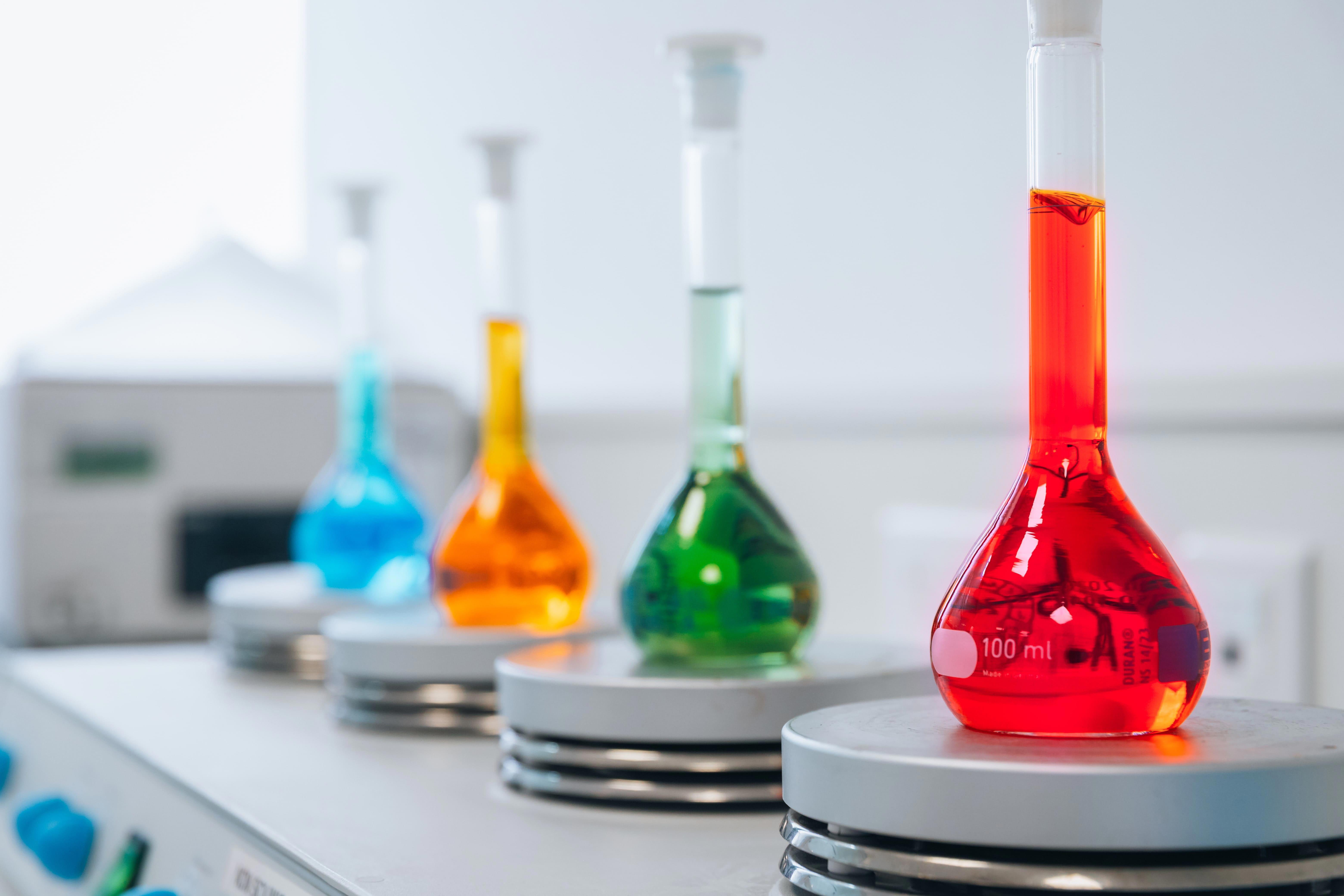 Quatre fioles Erlenmeyer contenant des solutions colorées (bleu, orange, vert et rouge) sont alignées sur des agitateurs magnétiques dans un laboratoire. La fiole au premier plan, graduée jusqu’à 100 ml, contient un liquide rouge. L’image illustre les activités de préparation et d’analyse de solutions réalisées par Eurofins BioPharma Product Testing dans le cadre du contrôle des résidus de stérilisation.