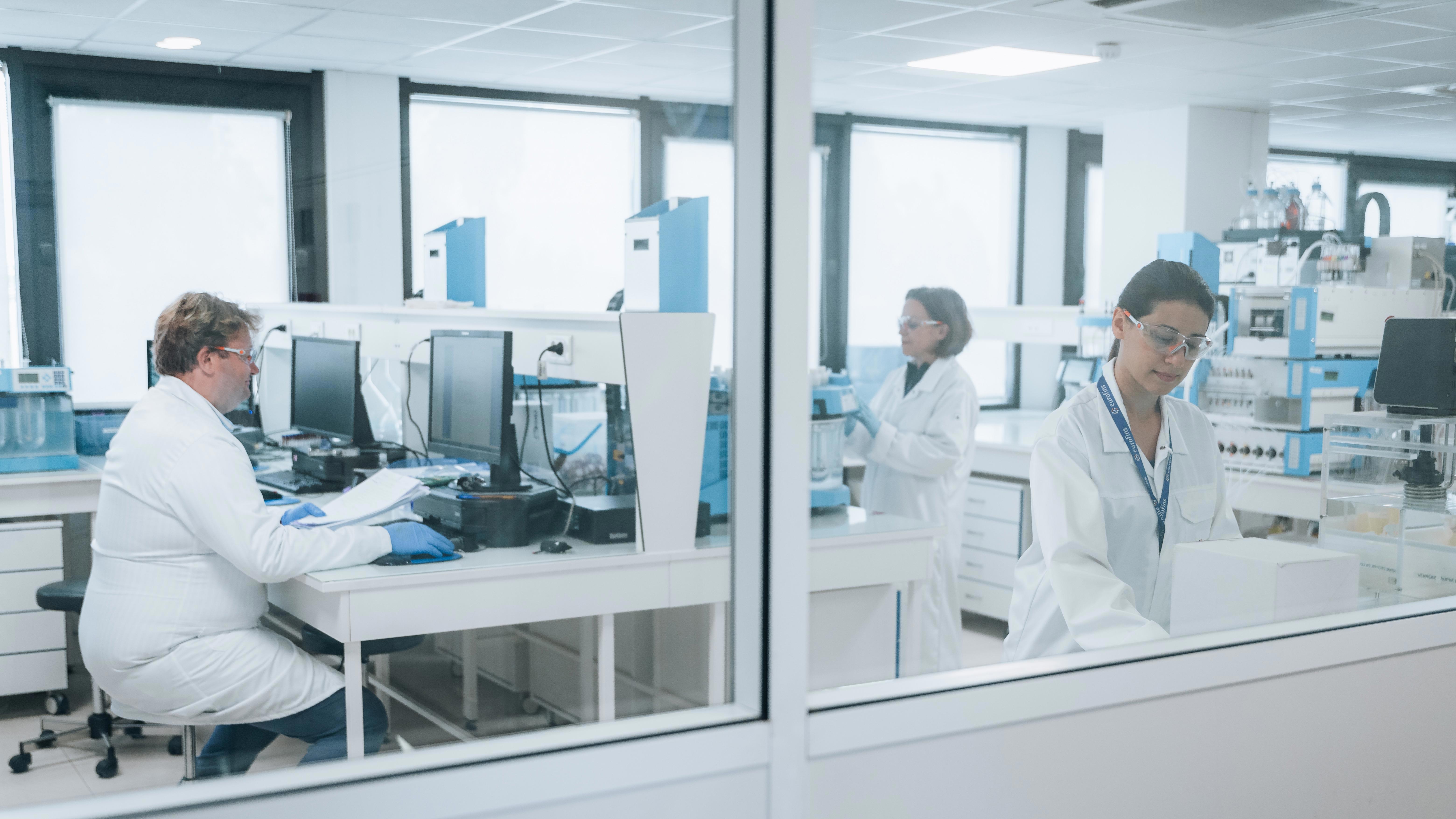 A modern laboratory with three individuals in white lab coats working on various tasks. One is seated at a desk with computer monitors, another is working with lab instruments in the background, and the third is focusing on equipment on the right.