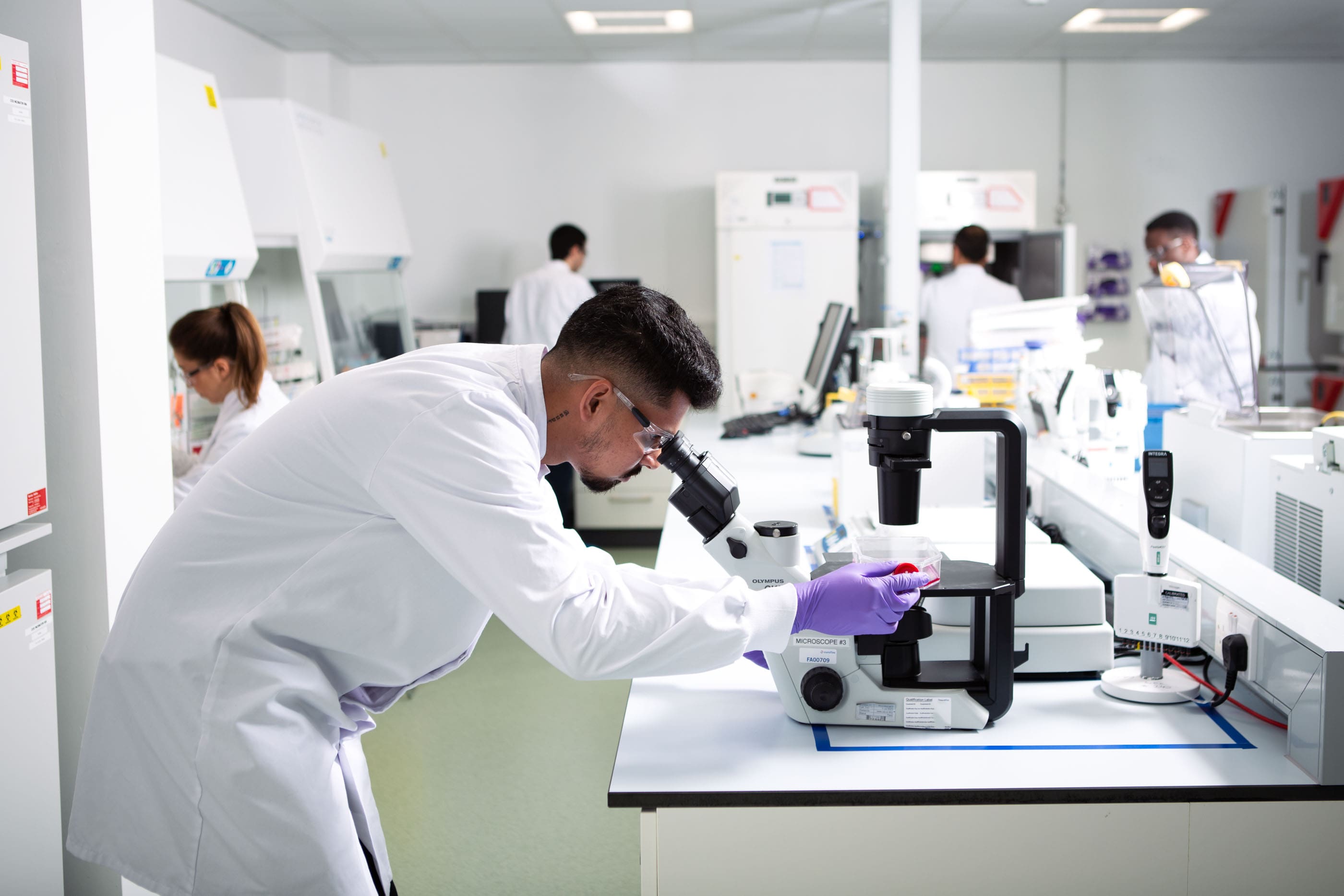 Scientifique en blouse blanche observant un échantillon au microscope dans un laboratoire Eurofins BioPharma Product Testing, équipé d’instruments analytiques modernes. L’image illustre les tests de compatibilité, de propreté et de performance réalisés sur les systèmes à usage unique et à usage multiple utilisés dans la fabrication et l’administration de produits biopharmaceutiques, conformément aux normes réglementaires internationales.