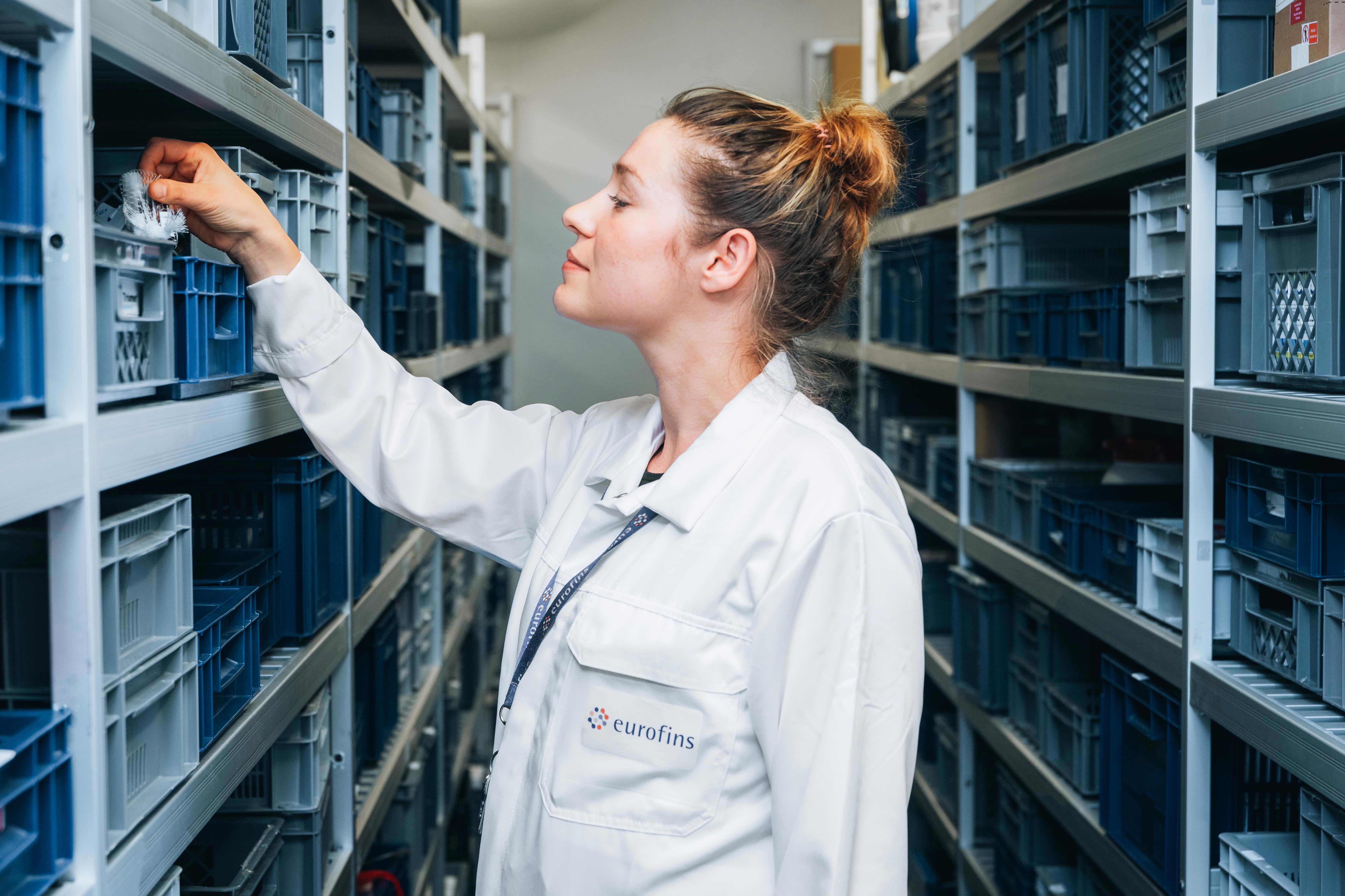 Technicienne de laboratoire Eurofins BioPharma Product Testing en blouse blanche, debout dans un espace de stockage organisé, entre deux rangées d’étagères métalliques contenant des bacs en plastique bleu et gris. Il tend la main pour saisir un objet dans un bac situé sur l’étagère de gauche.