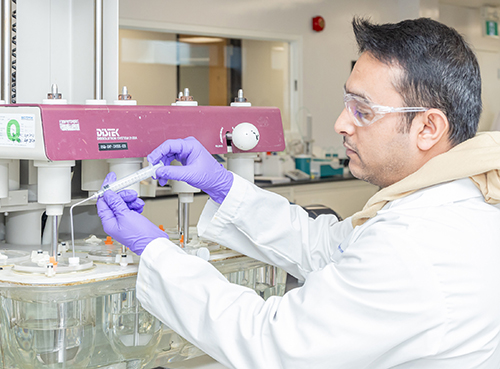 Scientifique en blouse de laboratoire, lunettes de protection et gants violets utilisant une pipette devant un appareil DISTEK équipé de récipients en verre contenant un liquide clair, illustrant les pratiques de laboratoire rigoureuses chez Eurofins BioPharma Product Testing.