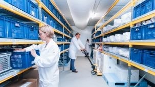  Zone de stockage chez Eurofins BioPharma Product Testing avec des étagères métalliques remplies de bacs bleus contenant des échantillons et du matériel de laboratoire. Des personnes en blouse blanche manipulent les bacs et utilisent un transpalette manuel pour déplacer des contenants, illustrant la gestion organisée des stocks et la conformité aux procédures de stabilité et de traçabilité.