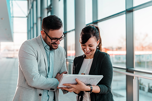 Deux professionnels en discussion dans un environnement de bureau moderne et lumineux, avec de grandes fenêtres. L’un tient une tablette ou un document que les deux examinent ensemble, illustrant une réunion ou une session de collaboration. L’image reflète les valeurs de communication, de travail d’équipe et de professionnalisme dans un contexte d’entreprise tel que celui d’Eurofins.