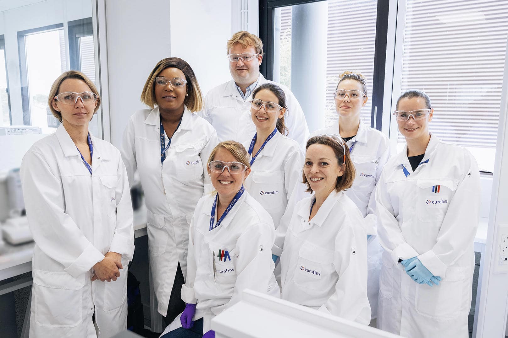 Groupe de collaborateurs Eurofins BioPharma Product Testing portant des blouses de laboratoire blanches et badges, réunis dans un environnement scientifique moderne. L’image montre un espace lumineux avec grandes fenêtres, mobilier technique et équipements analytiques, illustrant le travail collaboratif, la rigueur des procédures et la conformité aux normes qualité dans le domaine pharmaceutique et biopharmaceutique.