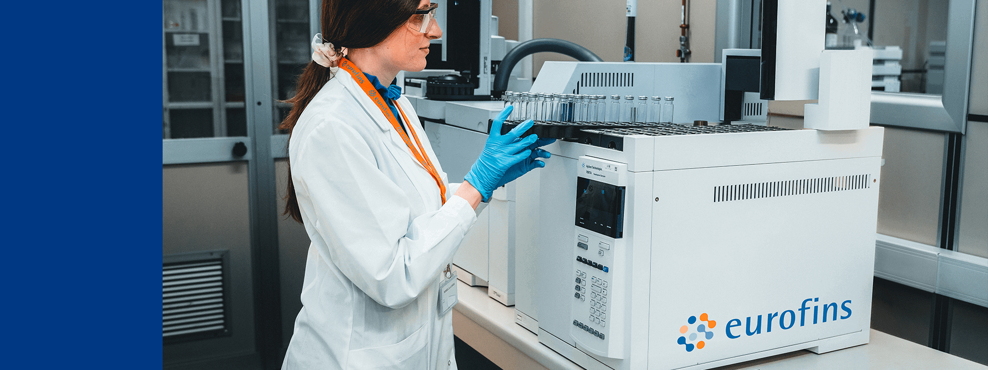 Dans un laboratoire Eurofins BioPharma Product Testing, un technicien en blouse blanche, gants bleus et lunettes de protection manipule un équipement analytique portant le logo Eurofins. Plusieurs flacons en verre sont disposés sur l’appareil, suggérant une opération de test pharmaceutique ou de contrôle qualité des gaz. À l’arrière-plan, on distingue des équipements scientifiques et des armoires de rangement, renforçant le cadre rigoureux et technique de l’environnement.