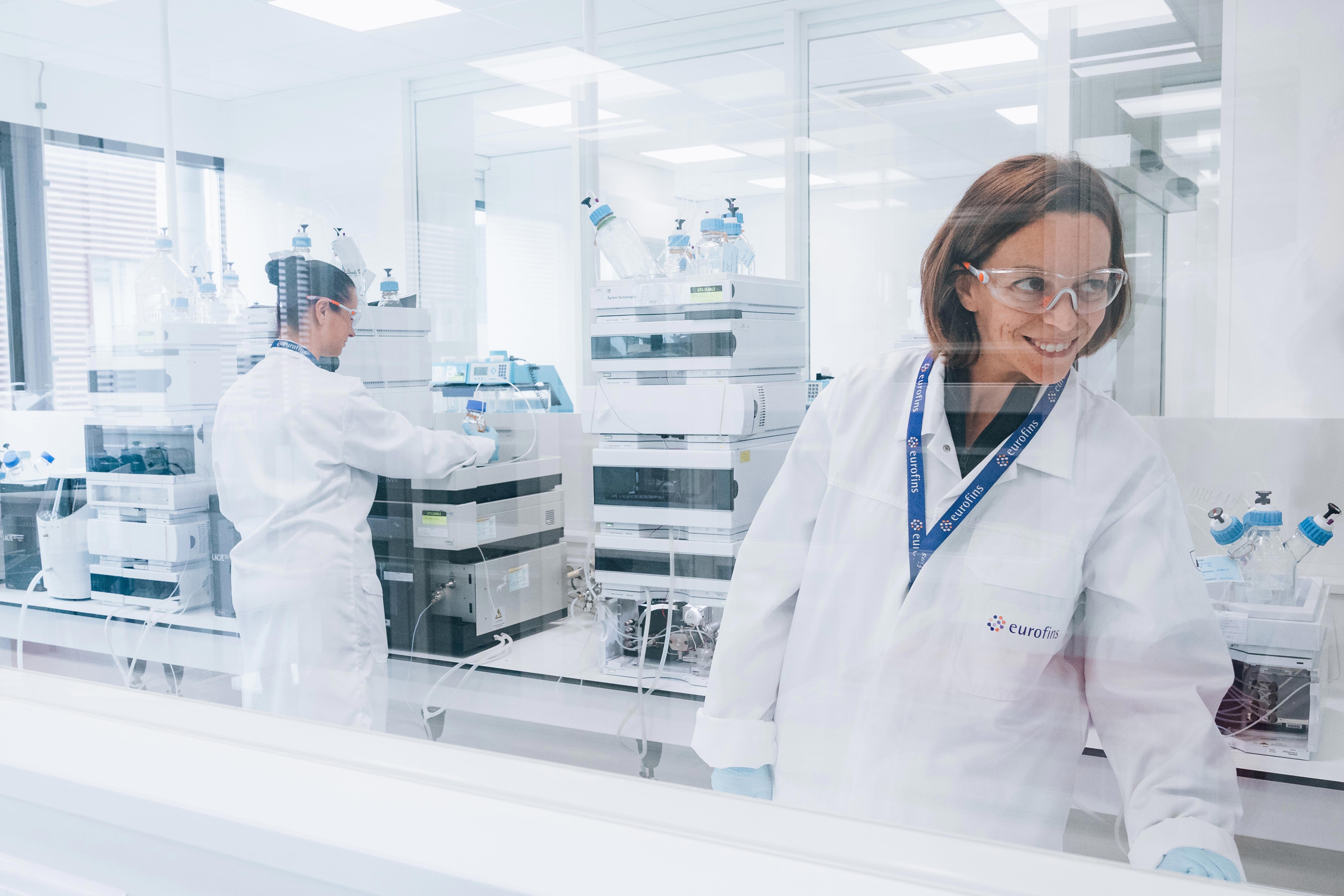 Deux collaborateurs d’Eurofins BioPharma Product Testing dans un laboratoire moderne, portant des blouses blanches, des gants et des lunettes de protection. L’un manipule un appareil scientifique tandis que l’autre regarde vers l’extérieur. En arrière-plan, divers équipements de laboratoire et bouteilles en verre avec bouchons bleus sont visibles dans un environnement propre et bien éclairé.
