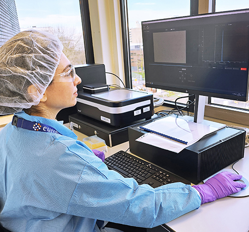 Technicien de laboratoire en blouse bleue, gants violets, filet pour cheveux et lunettes de protection, travaillant sur un ordinateur à double écran dans un environnement lumineux. Des équipements de laboratoire et des documents sont disposés sur le bureau. Cette image illustre les activités numériques et analytiques menées par Eurofins BioPharma Product Testing dans le cadre de ses opérations scientifiques.