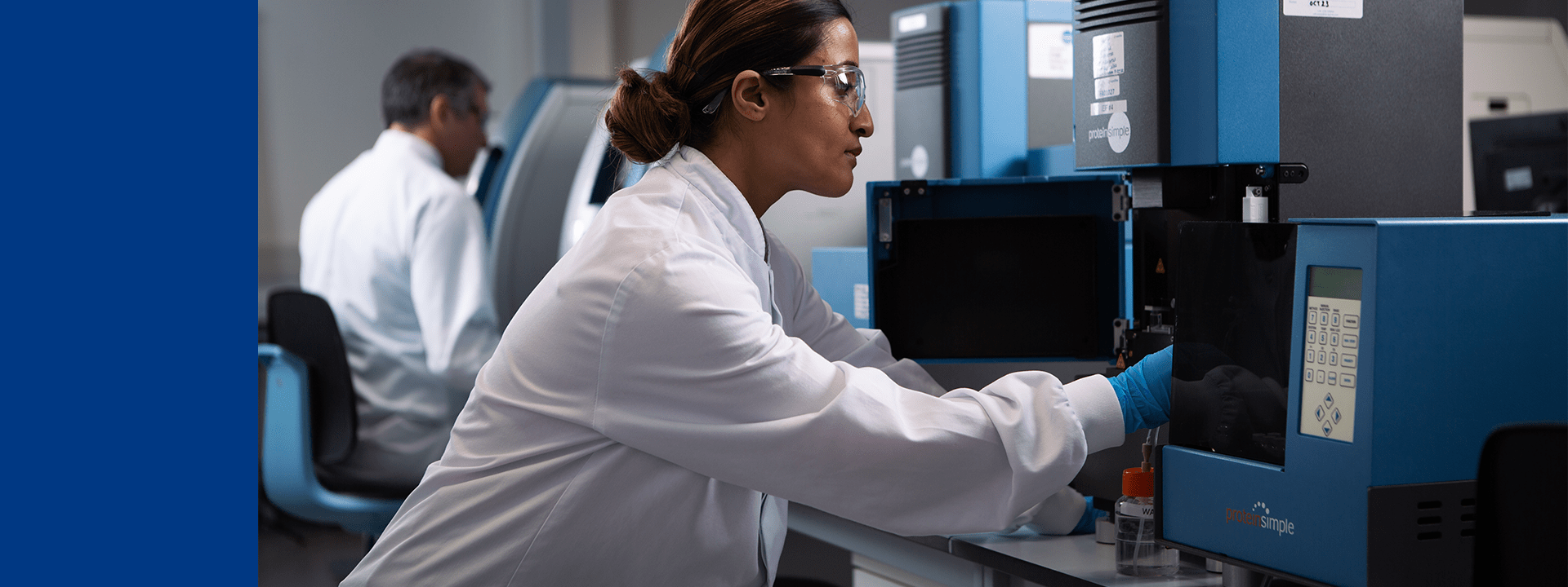 Technicienne de laboratoire Eurofins Biopharma Product Testing manipulant un instrument analytique de couleur bleue pour des tests en environnement contrôlé. L’image montre un espace de laboratoire moderne avec plusieurs équipements scientifiques, dont des systèmes analytiques et des postes de travail en arrière-plan.