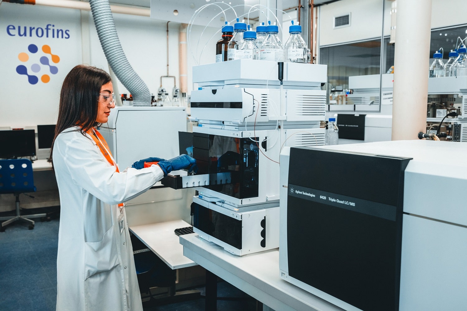 Laboratoire Eurofins BioPharma Product Testing équipé pour des analyses chromatographiques avancées. Une personne en blouse blanche et gants de protection manipule un système LC-MS (chromatographie liquide couplée à la spectrométrie de masse) pour le contrôle qualité et la détection d’impuretés. Sur l’appareil, plusieurs flacons de solvants sont disposés, et en arrière-plan, le logo Eurofins est visible sur le mur, illustrant un environnement conforme aux normes GMP.