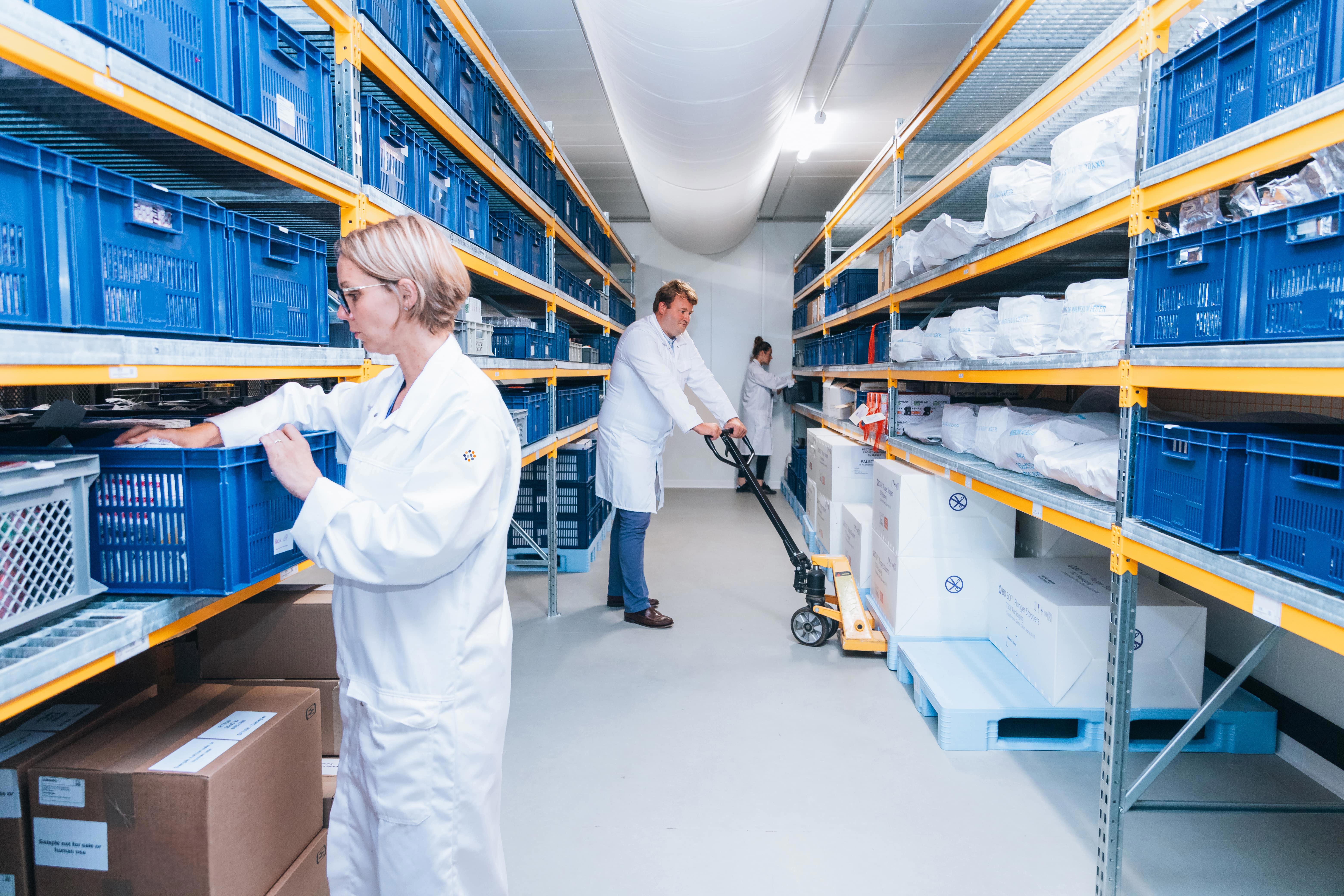 Trois collaborateurs d’Eurofins BioPharma Product Testing en blouse blanche travaillent dans un entrepôt de stockage. L’un manipule des objets dans un bac en plastique bleu, un autre utilise un transpalette pour déplacer des cartons, tandis qu’un troisième est visible au fond de l’allée. Les étagères métalliques jaunes et grises sont bien organisées et remplies de bacs bleus.