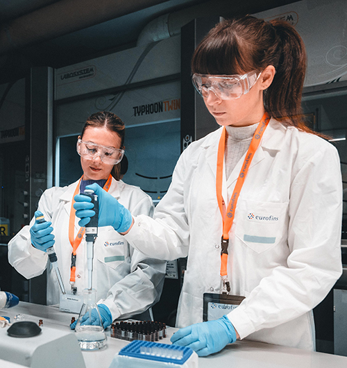 Deux techniciens de laboratoire chez Eurofins, portant des blouses blanches avec le logo Eurofins, des gants bleus, des lunettes de protection et des badges orange, manipulent des micropipettes dans un laboratoire moderne. L’environnement est équipé d’instruments scientifiques variés, illustrant les pratiques de précision, de sécurité et de conformité dans les analyses pharmaceutiques et bioanalytiques.