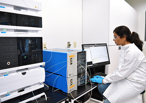 Scientifique en blouse blanche et gants bleus assis devant un ordinateur dans un laboratoire équipé d’instruments analytiques tels que des systèmes de chromatographie ou de spectrométrie de masse. L’écran affiche des données ou des graphiques. Cette image illustre les capacités d’analyse avancée d’Eurofins BioPharma Product Testing, notamment dans le cadre de la caractérisation de l’ARNm par SEC-MALS.