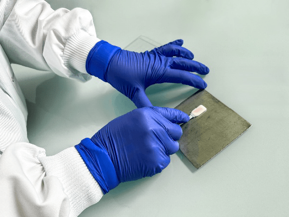 Technicien de laboratoire d’Eurofins BioPharma Product Testing manipulant un échantillon à l’aide d’une pince sur une plaque métallique, avec des gants en nitrile bleus et une blouse blanche, dans le cadre d’un processus de développement et validation du nettoyage.