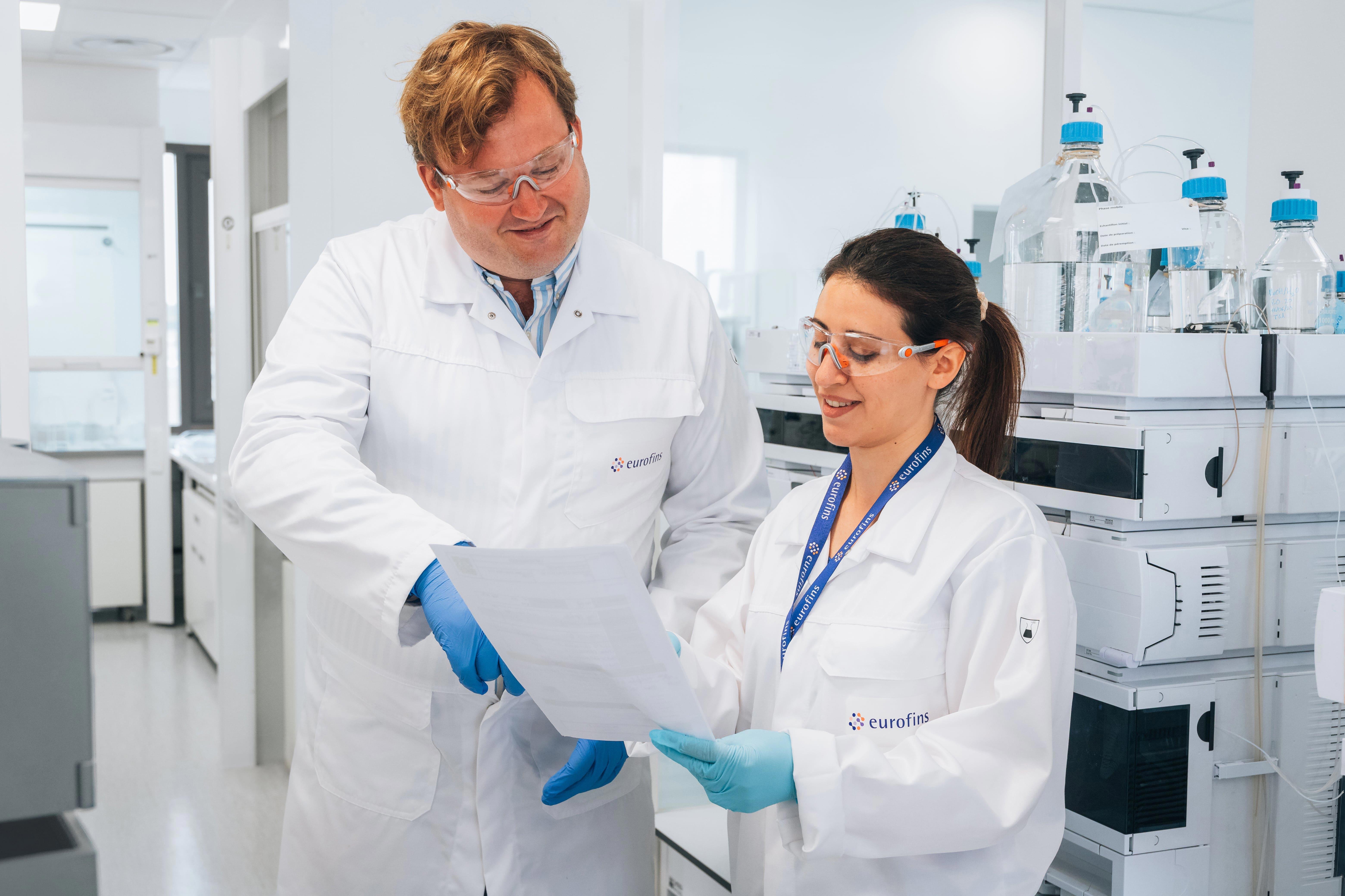 Deux analystes en laboratoire, vêtus de blouses blanches et de lunettes de protection, examinent un document technique dans un environnement de laboratoire moderne. L’un des collaborateurs porte des gants nitrile bleus. En arrière-plan, des équipements analytiques et des flacons en verre avec bouchons bleus sont visibles. L’image illustre les essais de dégradations forcées réalisés par Eurofins BioPharma Product Testing dans le cadre de la caractérisation de la stabilité des produits pharmaceutiques.