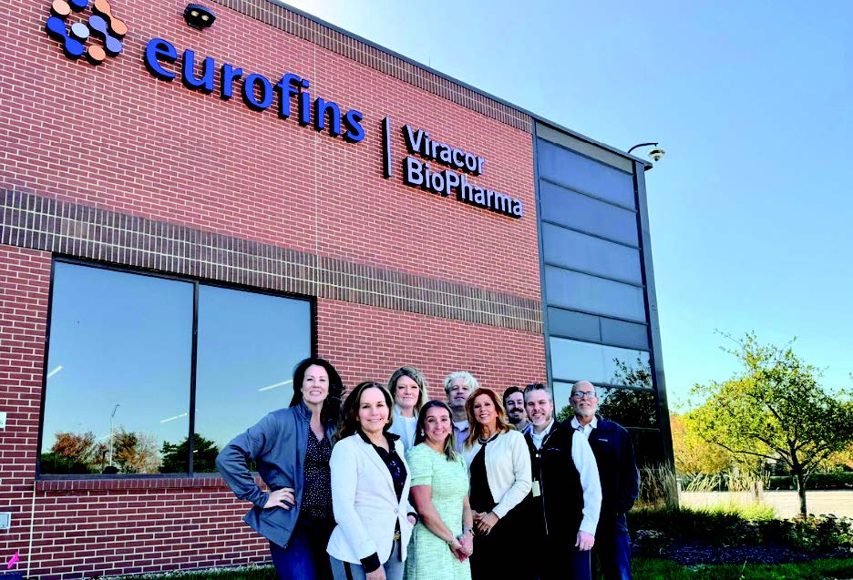 Groupe de collaborateurs Eurofins devant le bâtiment Eurofins Viracor BioPharma. L’image montre une façade en briques rouges avec le logo Eurofins et Viracor BioPharma, de grandes fenêtres réfléchissant le ciel bleu et des arbres en arrière-plan.