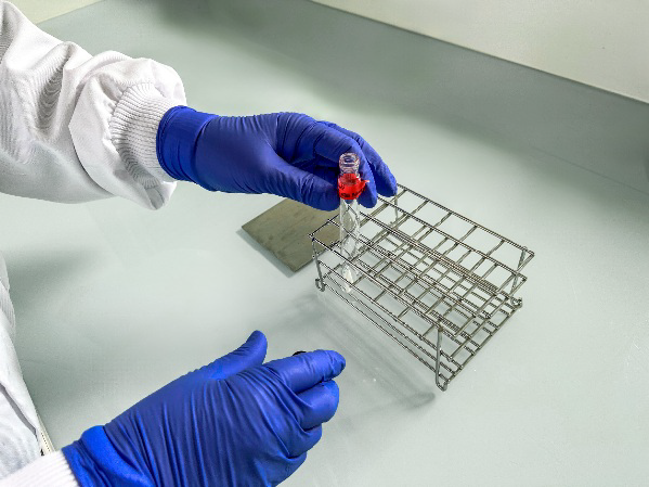 Technicien de laboratoire d’Eurofins BioPharma Product Testing manipulant un tube à essai contenant un liquide rouge, à l’aide de gants bleus. Le tube est placé dans un support métallique sur une surface de travail propre, dans le cadre du développement et de la validation du nettoyage.