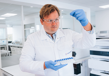 Close-up of a laboratory professional wearing a white lab coat, safety glasses, and blue gloves holding up a clear vial for inspection, with a rack of small sample tubes aligned on a work surface in a bright, modern laboratory.