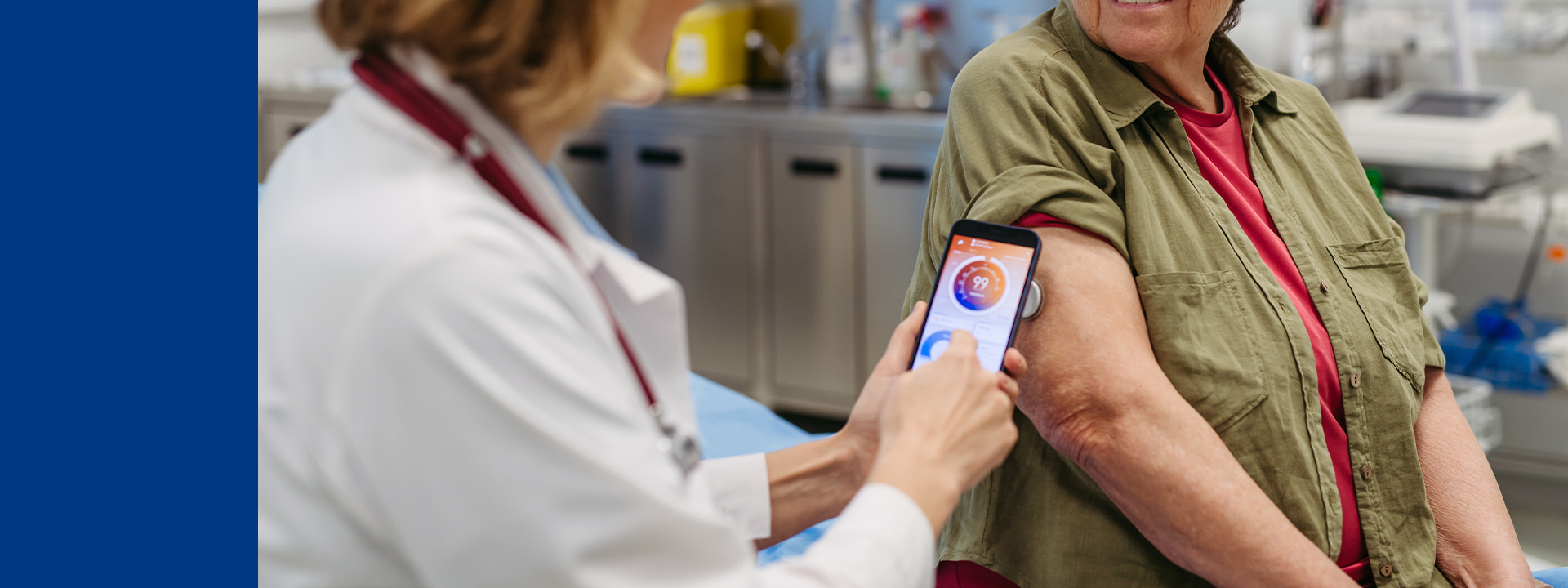 Healthcare professional showing a mobile device displaying a heart monitoring app to an elderly patient in a clinical setting, with medical equipment visible in the background.
