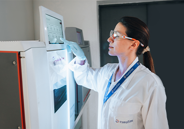 Laboratory professional wearing a white lab coat, gloves, and safety glasses operating a laboratory instrument by touching a digital control panel, in a clean, well-lit lab environment.