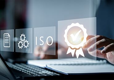 Close-up of a laptop on a desk with overlaid digital interface graphics, including the text “ISO,” gear icons, a document symbol, and a certification badge with a checkmark, representing quality standards, compliance, and certification processes.