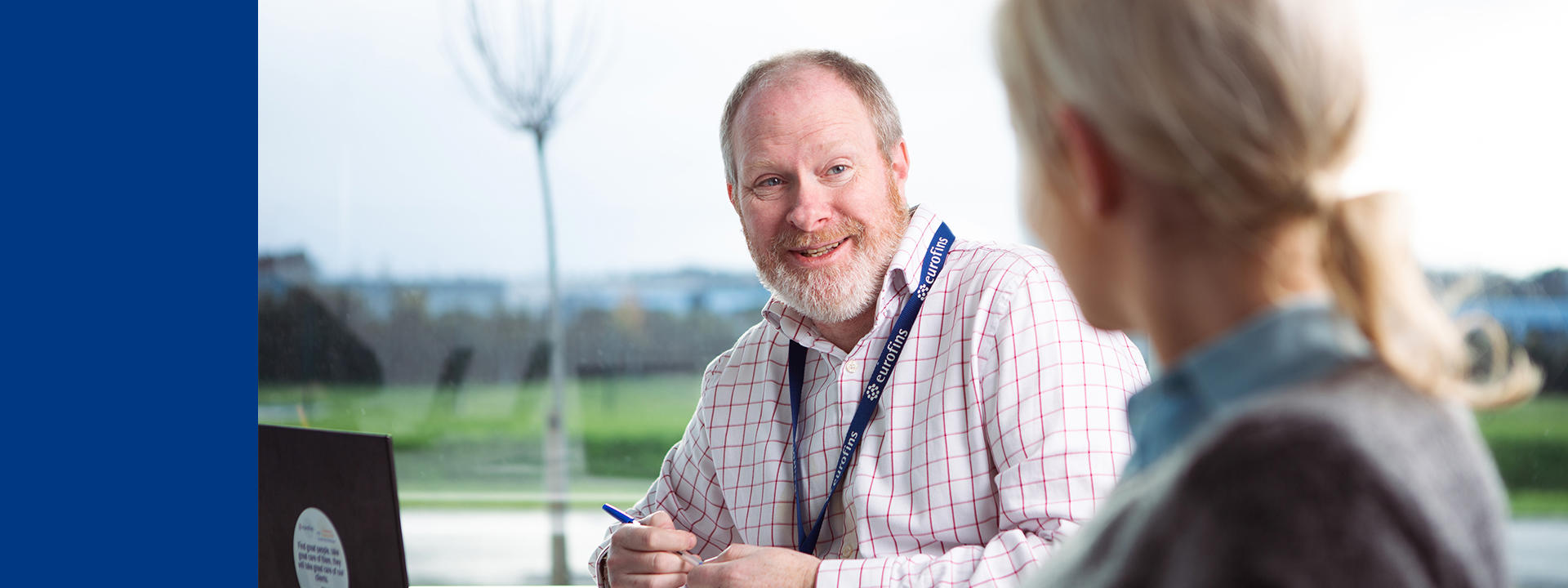 Two people seated at an outdoor table, engaged in conversation, with one reviewing a laptop while the other listens, and a soft, natural landscape visible in the background.