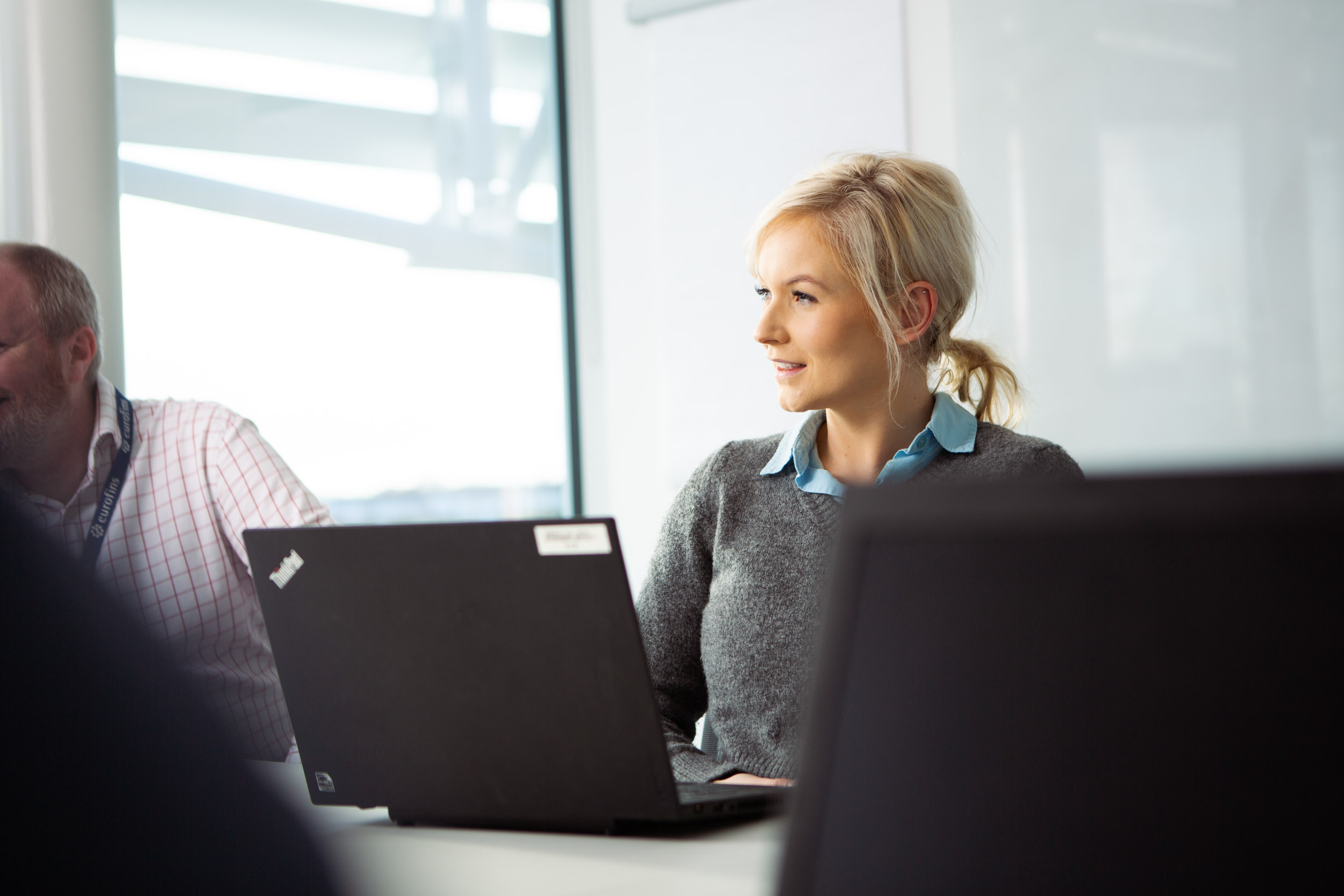 Professional seated at a table in a modern office, working on a laptop during a meeting, with other participants partially visible and soft lighting from nearby windows.