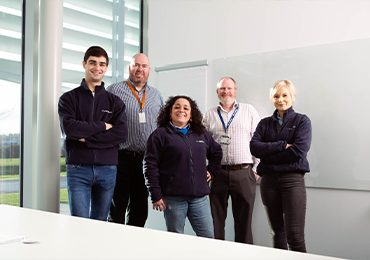 Group of five professionals standing together in a bright office space with large windows and a whiteboard, dressed in business-casual attire, positioned in a row with arms crossed behind a conference table.