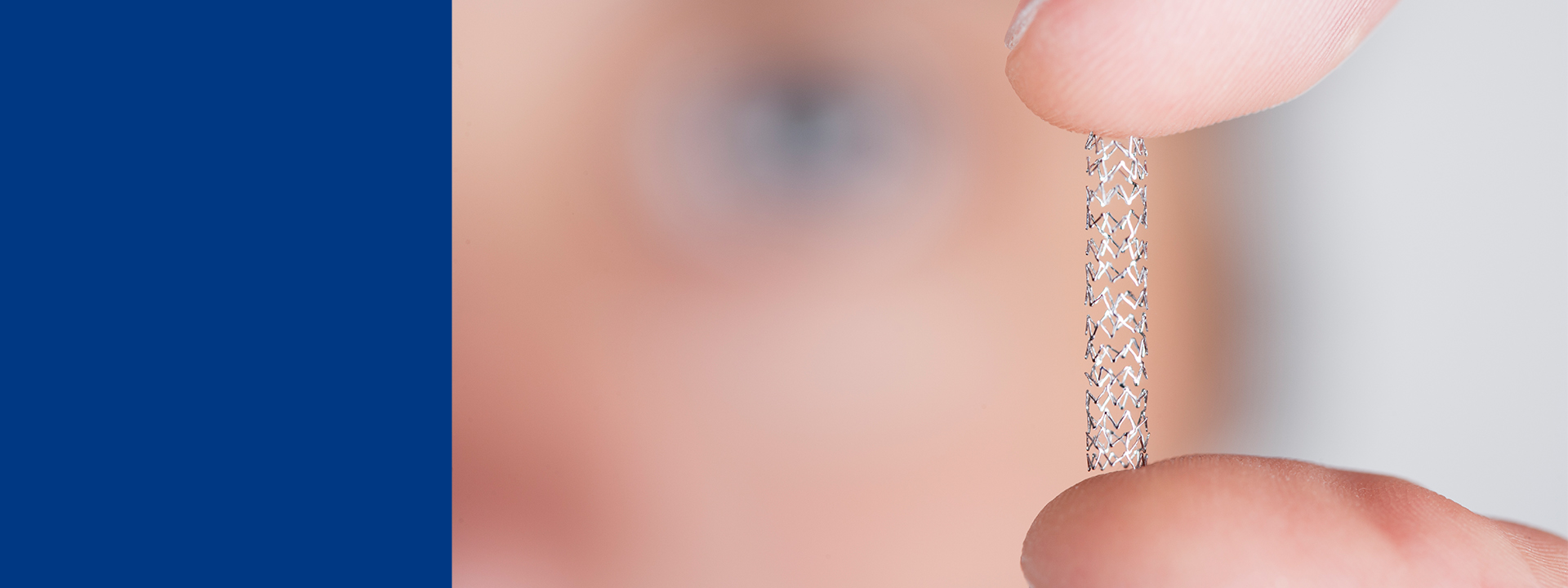 Close-up of fingers holding a small cylindrical implantable medical device with a mesh-like structure, shown against a blurred background, illustrating miniature device design and precision engineering.