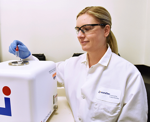 Laboratory professional wearing safety glasses, a white lab coat, and blue gloves placing a small sample onto a compact analytical instrument, with the Eurofins logo visible on the coat in a clean lab setting.