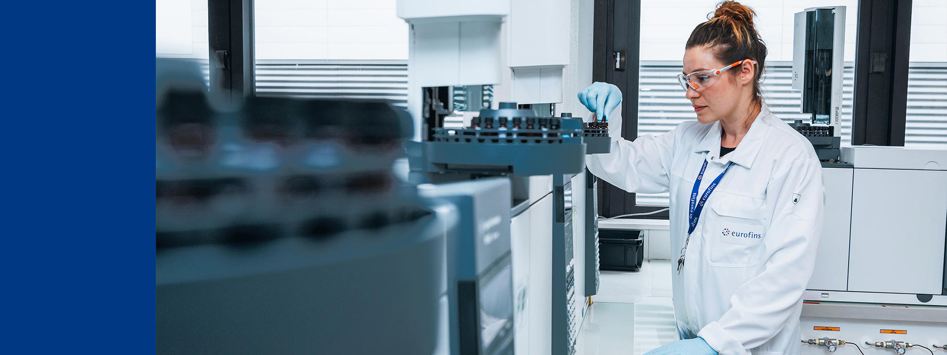 Laboratory professional in a white lab coat and safety glasses standing beside automated analytical instruments, monitoring equipment in a bright, modern laboratory with rows of machinery in the foreground.