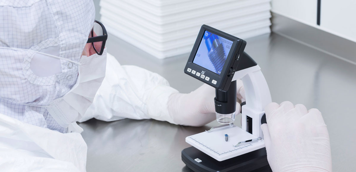 Laboratory professional in full cleanroom attire using a microscope with a digital display to examine a sample on a workbench in a controlled lab environment.