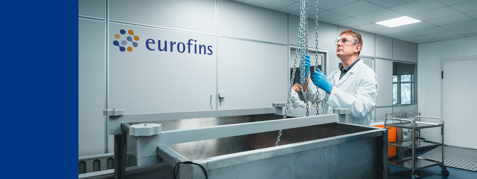 Laboratory professional wearing a white lab coat, safety glasses, and gloves lifting a chain mechanism above a large stainless steel tank in a clean industrial laboratory, with the Eurofins logo visible on the wall in the background.