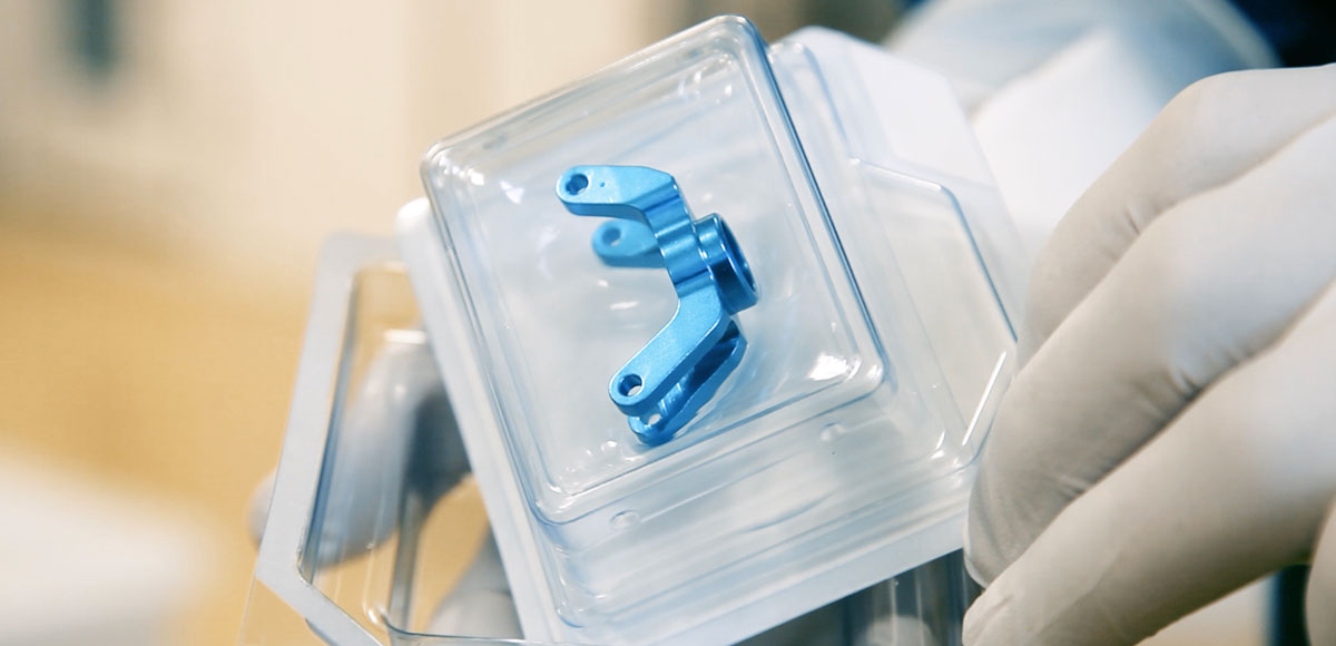 Blue metallic medical device with two arms, each with an opening at the end, in a sealed blister pack.