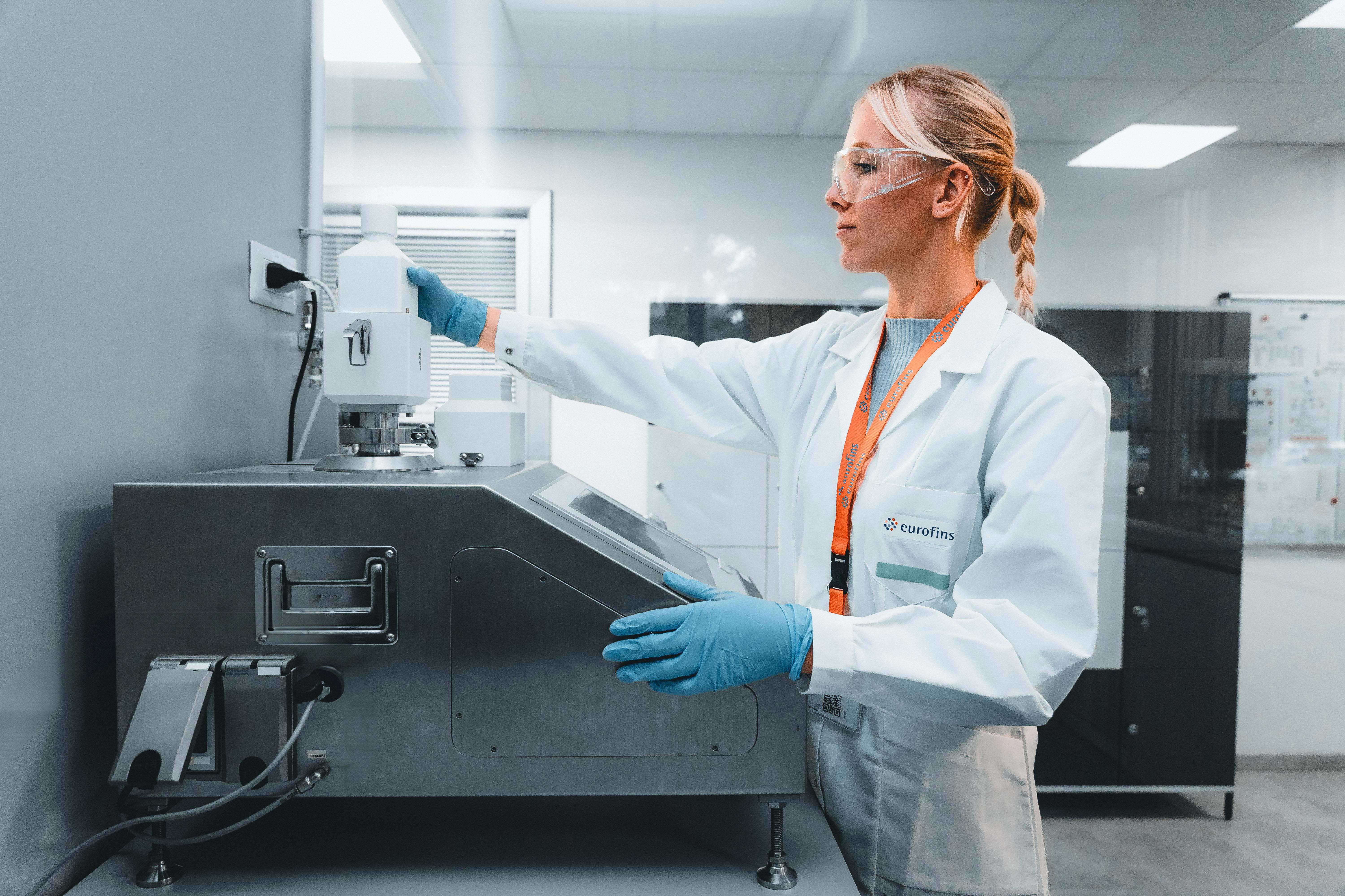 Laboratory professional wearing a white lab coat, blue gloves, and safety glasses standing beside a stainless steel work surface, reaching up to operate a wall-mounted lab instrument in a clean, well-lit laboratory environment.