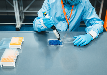 Laboratory professional wearing cleanroom attire and blue gloves using a pipette to transfer liquid into a multi‑well plate on a workbench, with additional sample trays arranged nearby in a controlled lab environment.