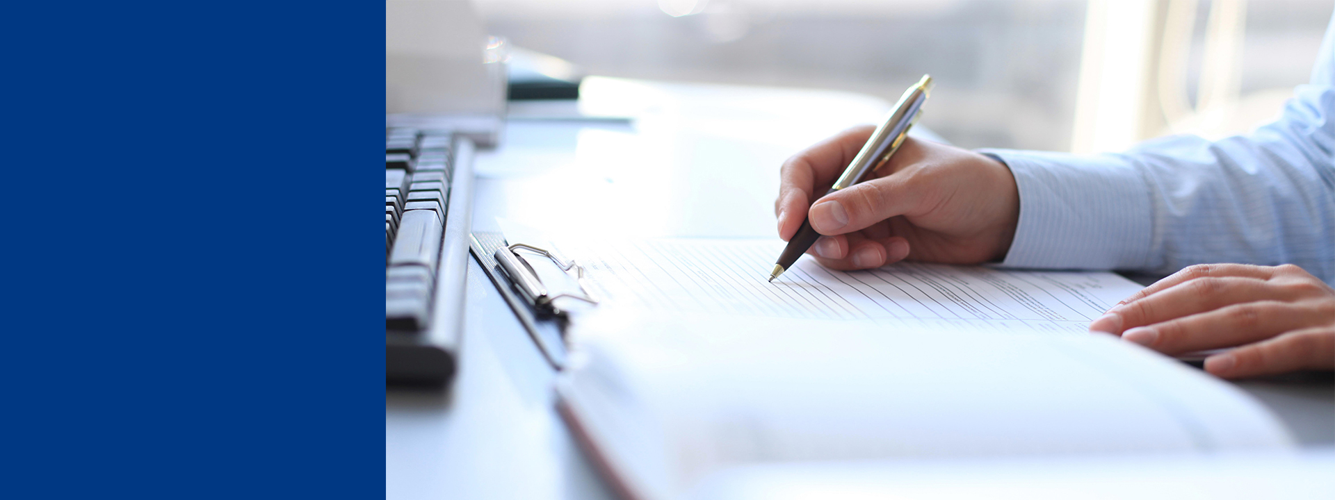 Close-up of a person’s hand writing on a document with a pen at a desk, with a computer keyboard and clipboard visible nearby, representing note-taking or document completion in a professional setting.