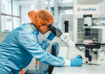 Laboratory professional in cleanroom attire, including a hair cover and gloves, using a microscope to examine a sample, with analytical equipment and the Eurofins logo visible in a bright laboratory setting.