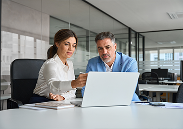 Two professionals seated at a desk in a modern office with glass walls, collaborating while reviewing content on a laptop, with a smartphone and documents visible on the workspace.