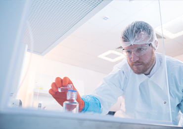 Close-up of a laboratory professional wearing protective gloves, lab coat, hair cover, and safety glasses, carefully holding a small vial of liquid. The individual is viewed through a transparent surface, with a softly lit laboratory environment and equipment visible in the background.
